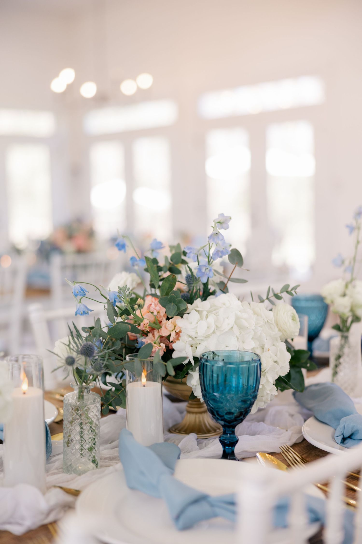 Elegant table settings with blue glassware, white flowers and candles in a bright reception space.
