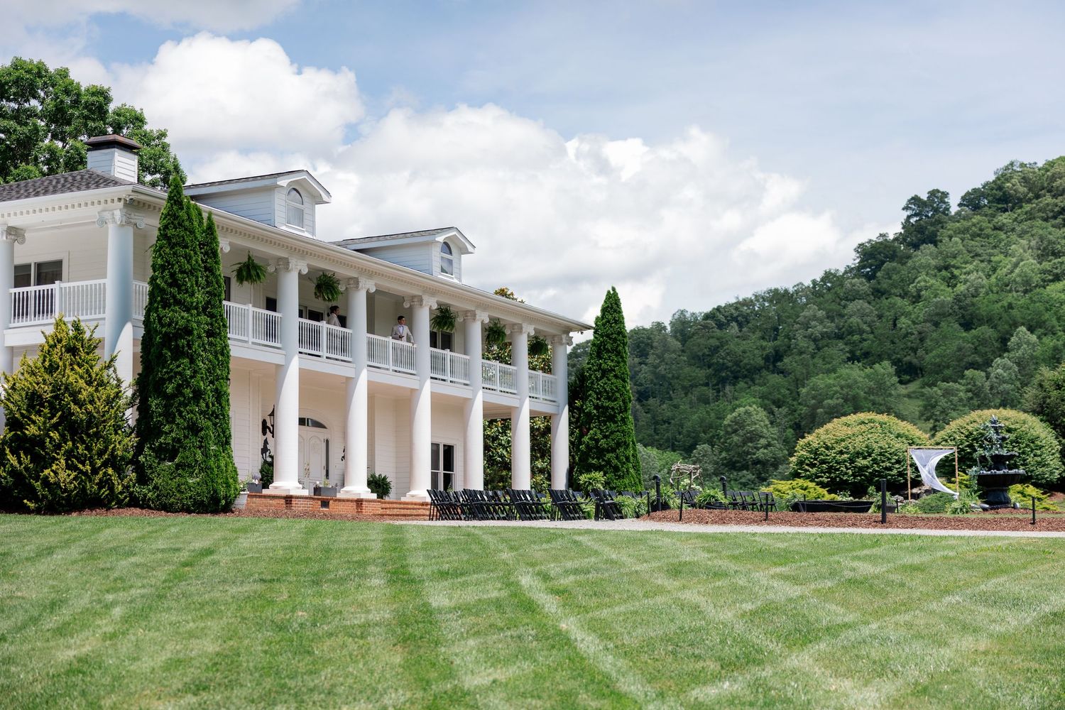 Elegant white mansion with tall pillars and manicured green lawns stretches across a panoramic landscape view.