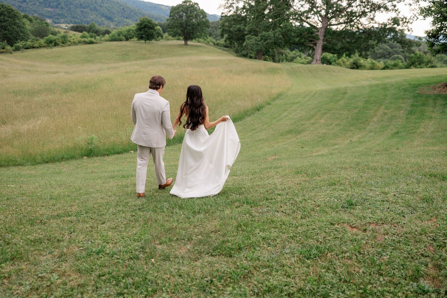 A couple walks hand in hand across a green hillside with mountains in the distance.