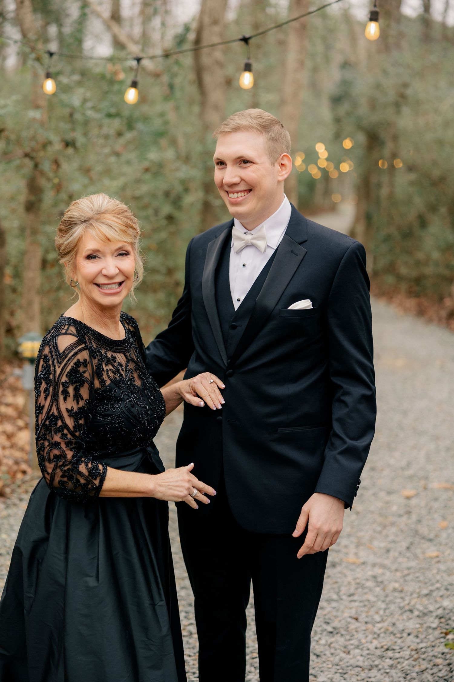 Outdoor wedding portrait with string lights and gravel path in wooded setting.
