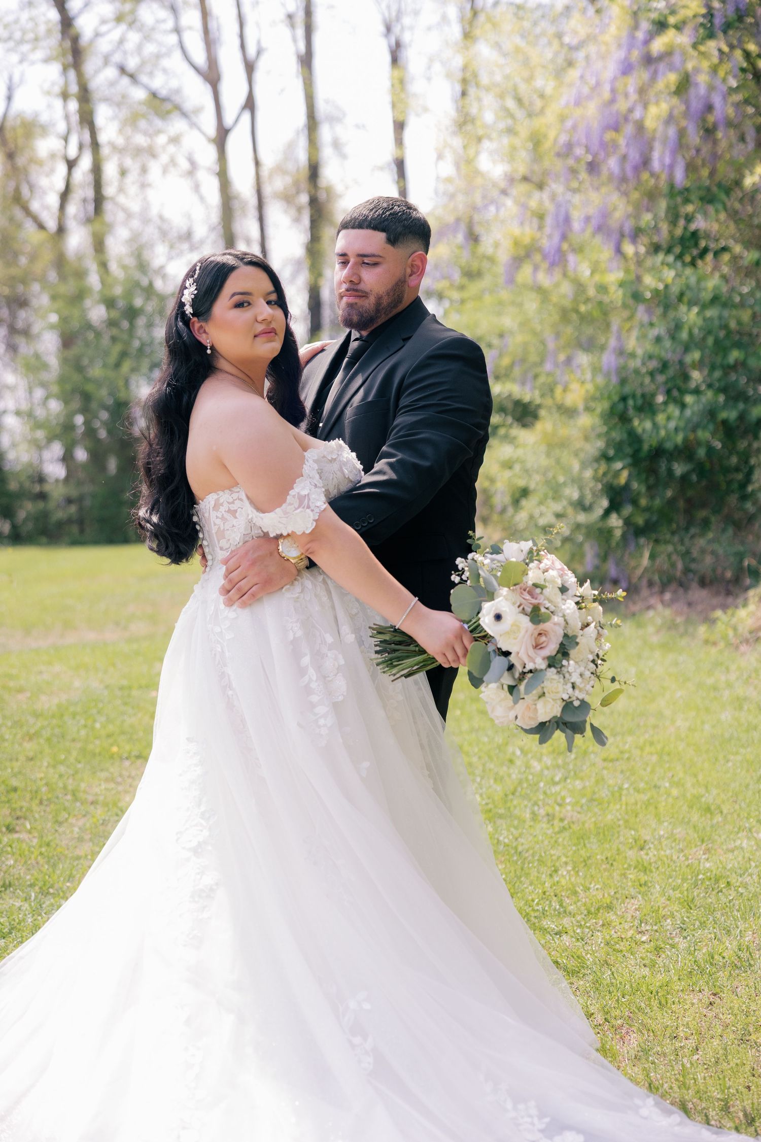 A romantic wedding portrait of a couple outdoors with wisteria blooms, the bride in a beautiful lace off-shoulder gown.