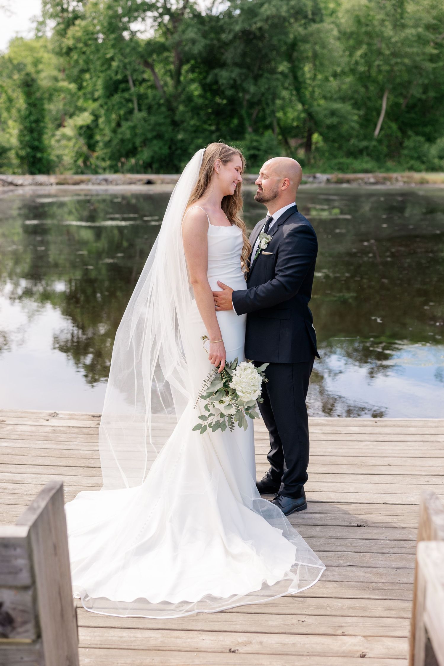 Romantic wedding couple sharing a moment on a wooden dock by a tranquil lake.