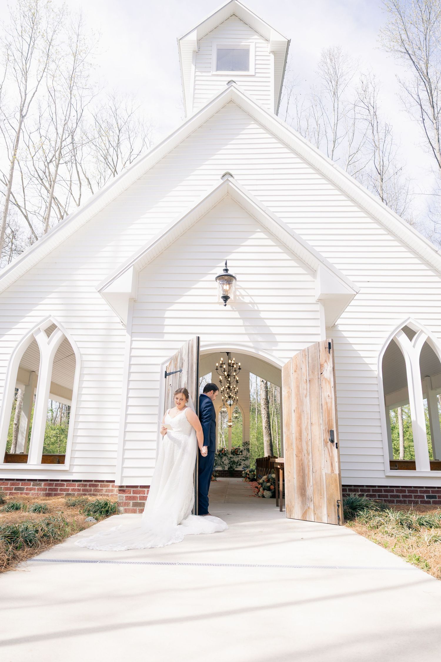 Wedding couple stands in doorway of charming white chapel with gothic windows and rustic wooden door.
