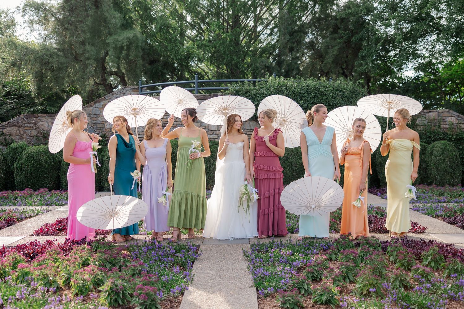 Bridesmaids in colorful dresses pose with white parasols in a garden setting.