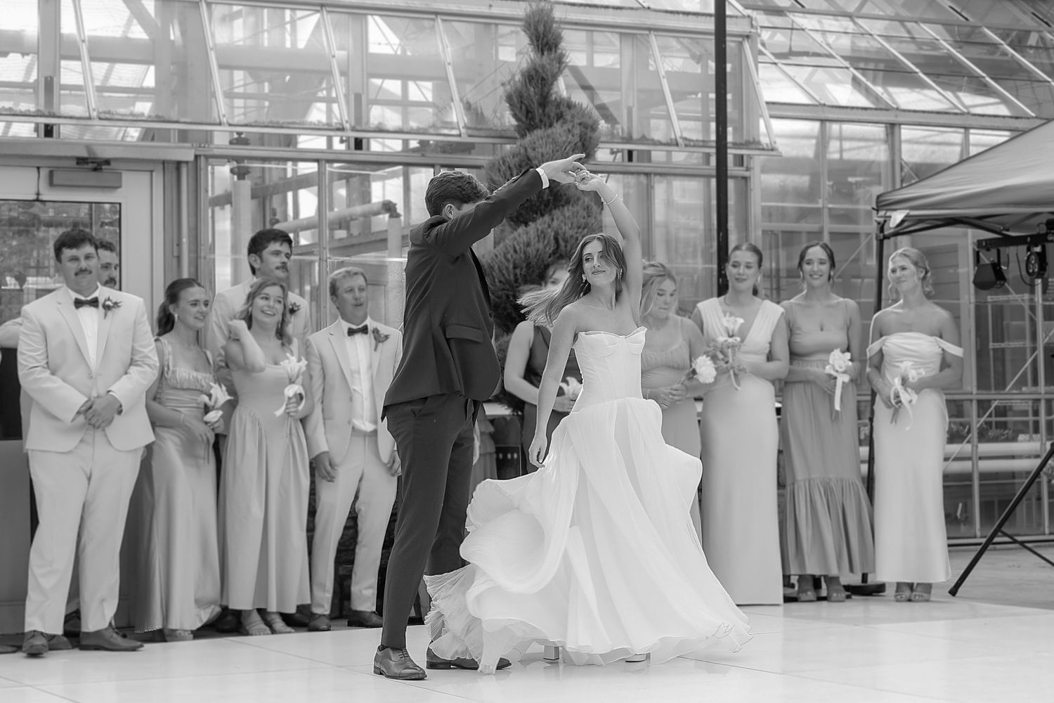 Black and white photo of wedding reception in greenhouse with seated guests and dancing couple.
