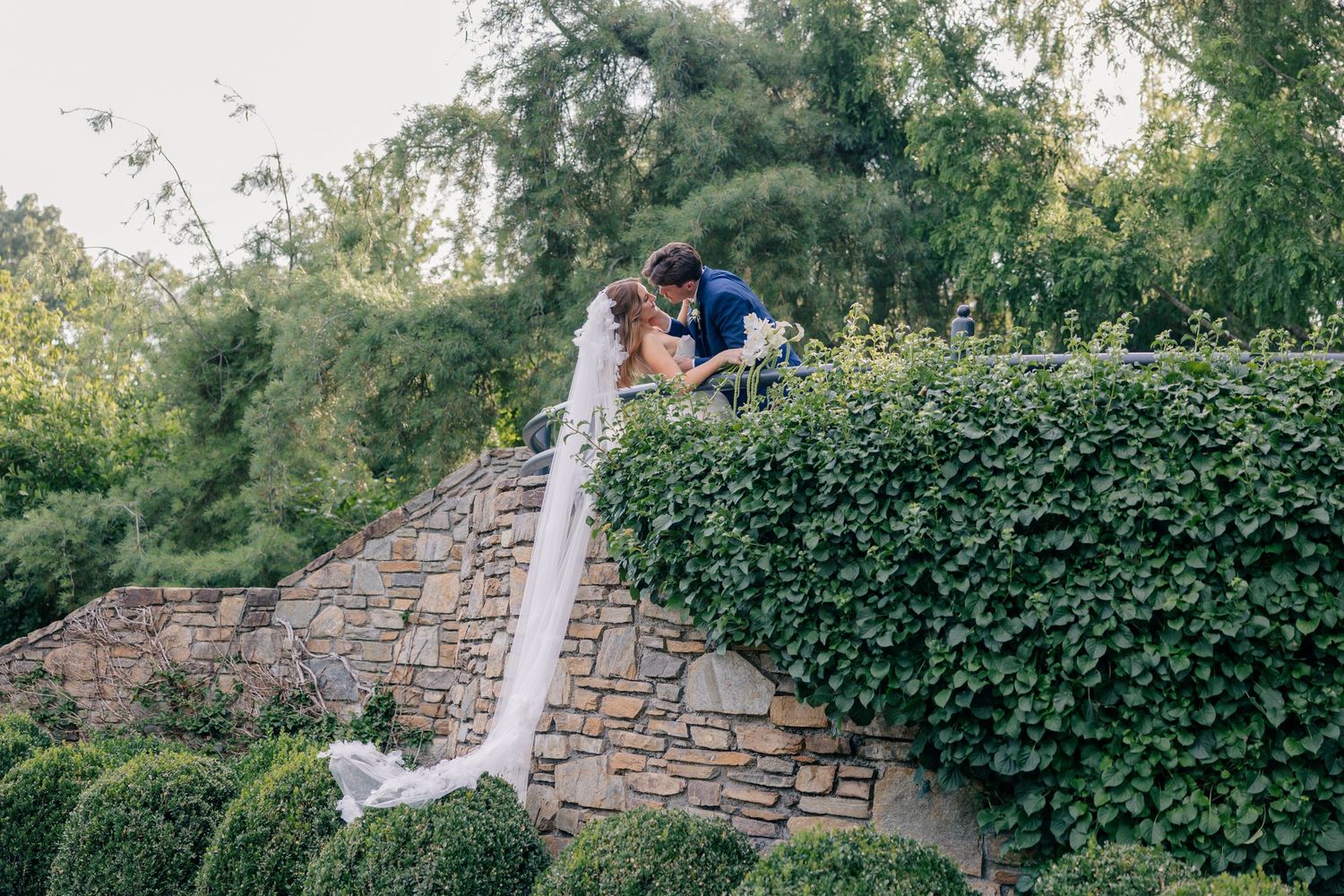 A romantic outdoor wedding ceremony takes place in a lush garden setting with stone walls and greenery.