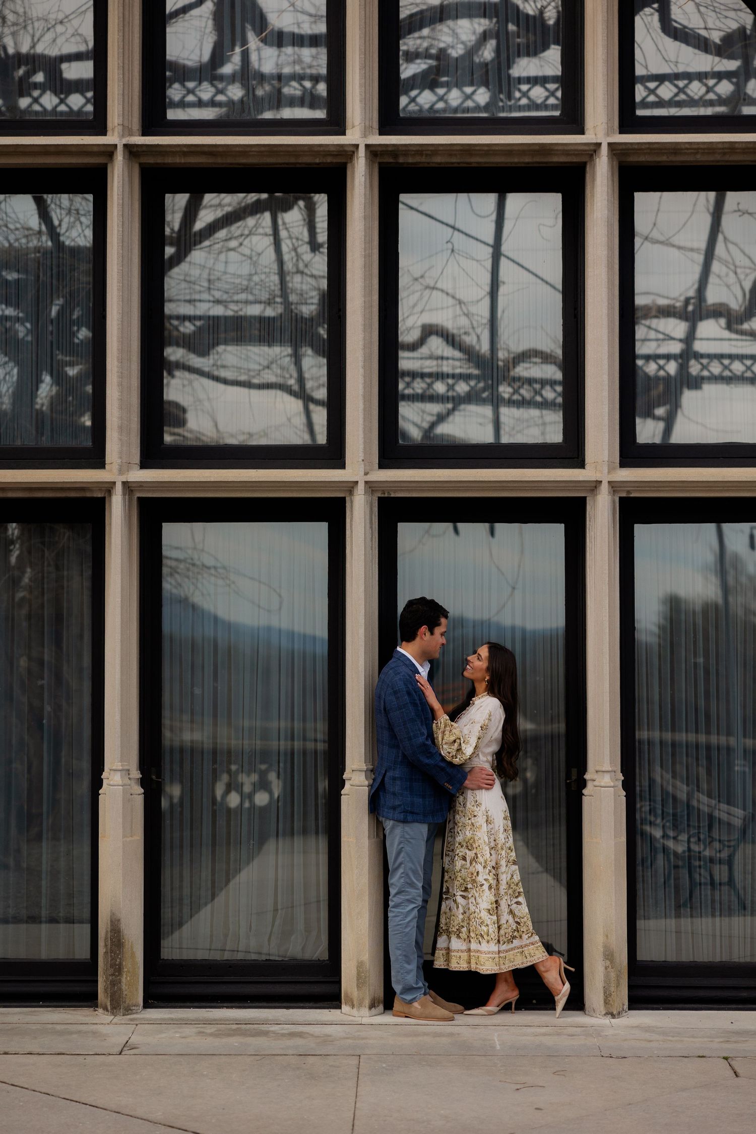 A couple embraces in front of a modern glass building facade with geometric window panels.