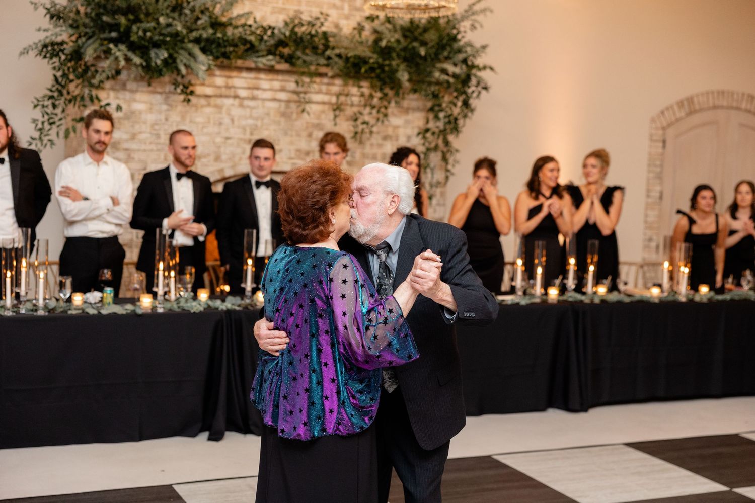 Older couple shares a dance at a wedding reception in front of a decorated head table with greenery and black linens.