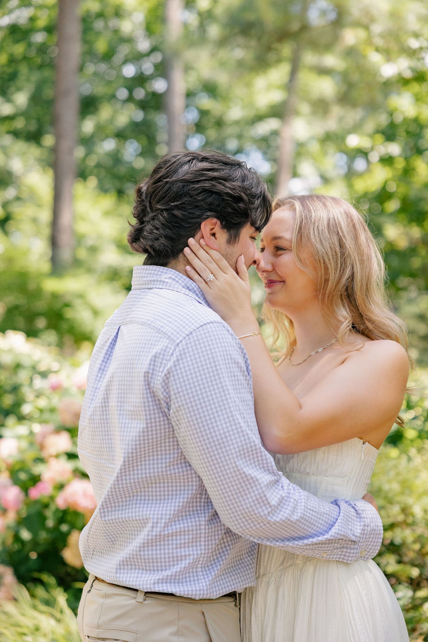 A couple shares a romantic almost-kiss moment outdoors, surrounded by lush green trees and colorful blooming flowers.