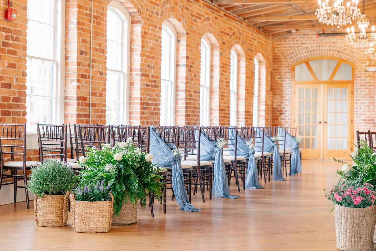 Elegant wedding venue with brick walls, arched windows, chiavari chairs with blue sashes, and floral arrangements in wicker baskets.