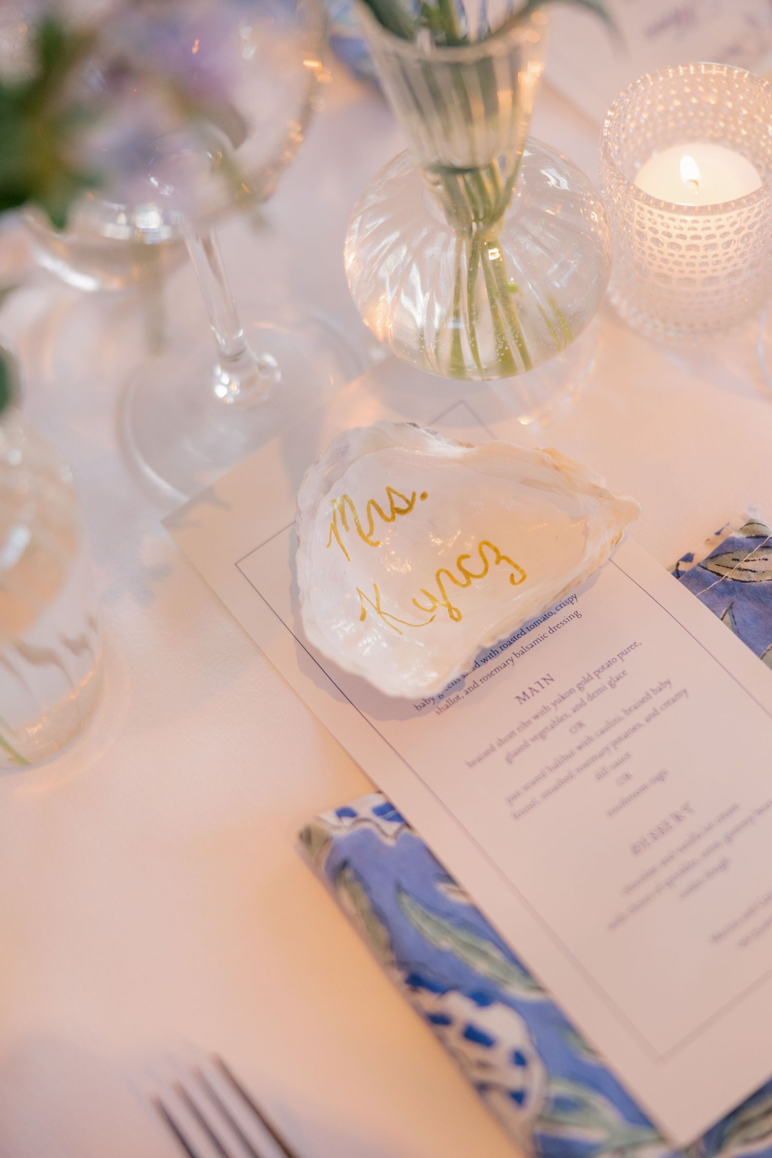 Elegant wedding place setting with oyster shell name card reading Mrs. Hynes, menu, and blue napkin.
