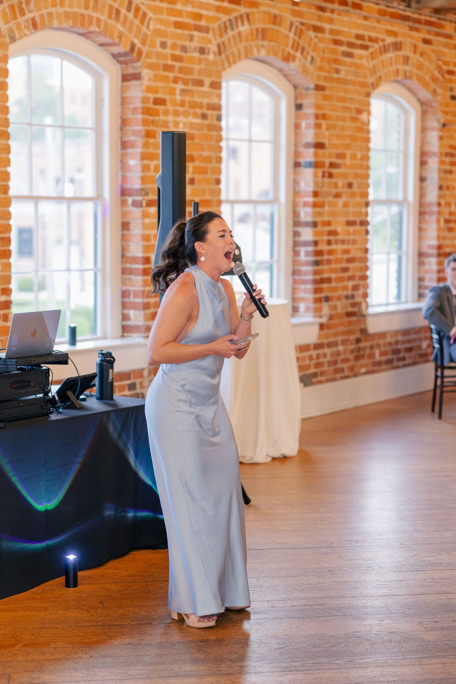 A woman in a silver gown delivers a heartfelt speech at a wedding reception in a brick venue.