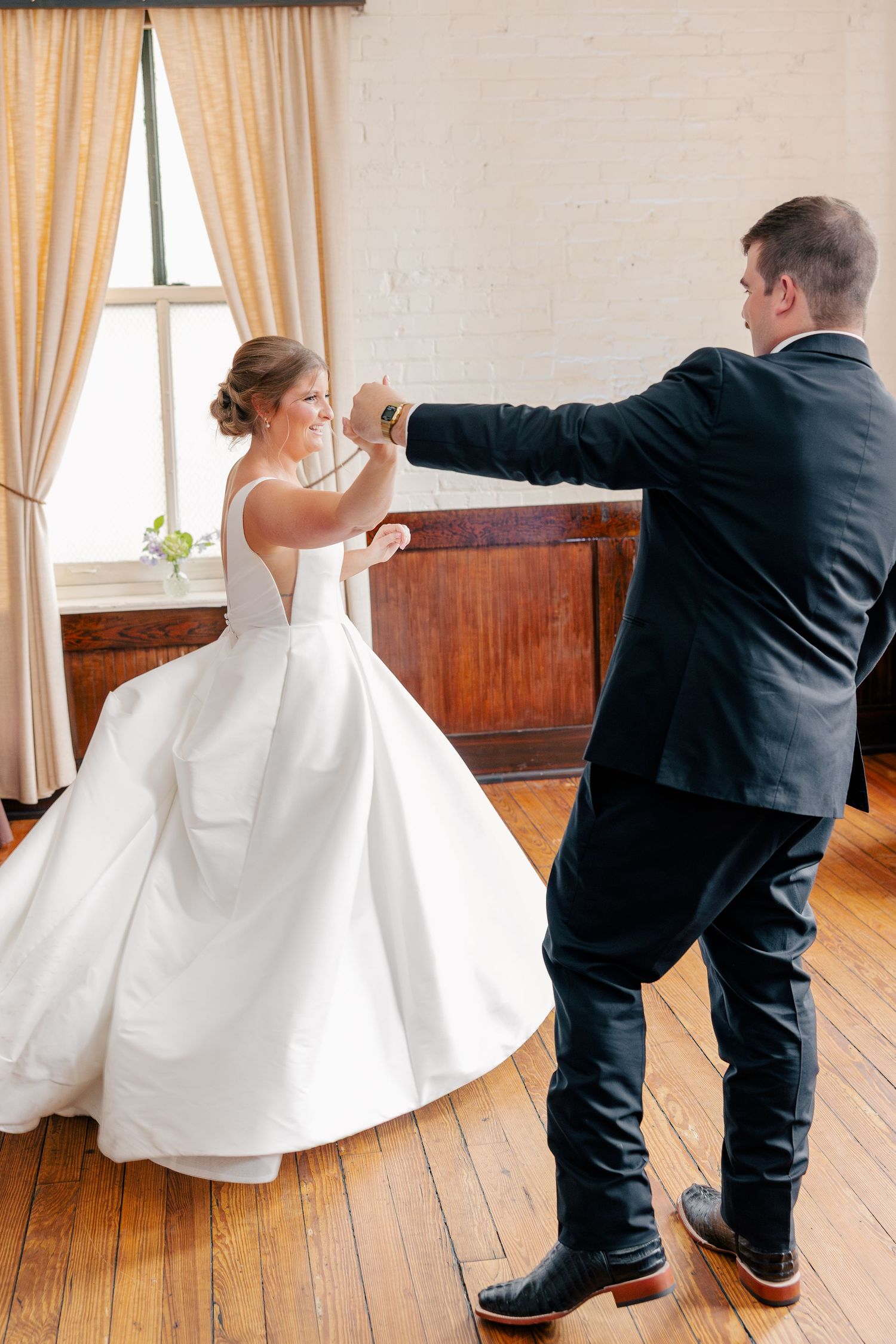 Groom twirls bride in elegant white satin gown during first dance in charming rustic venue.