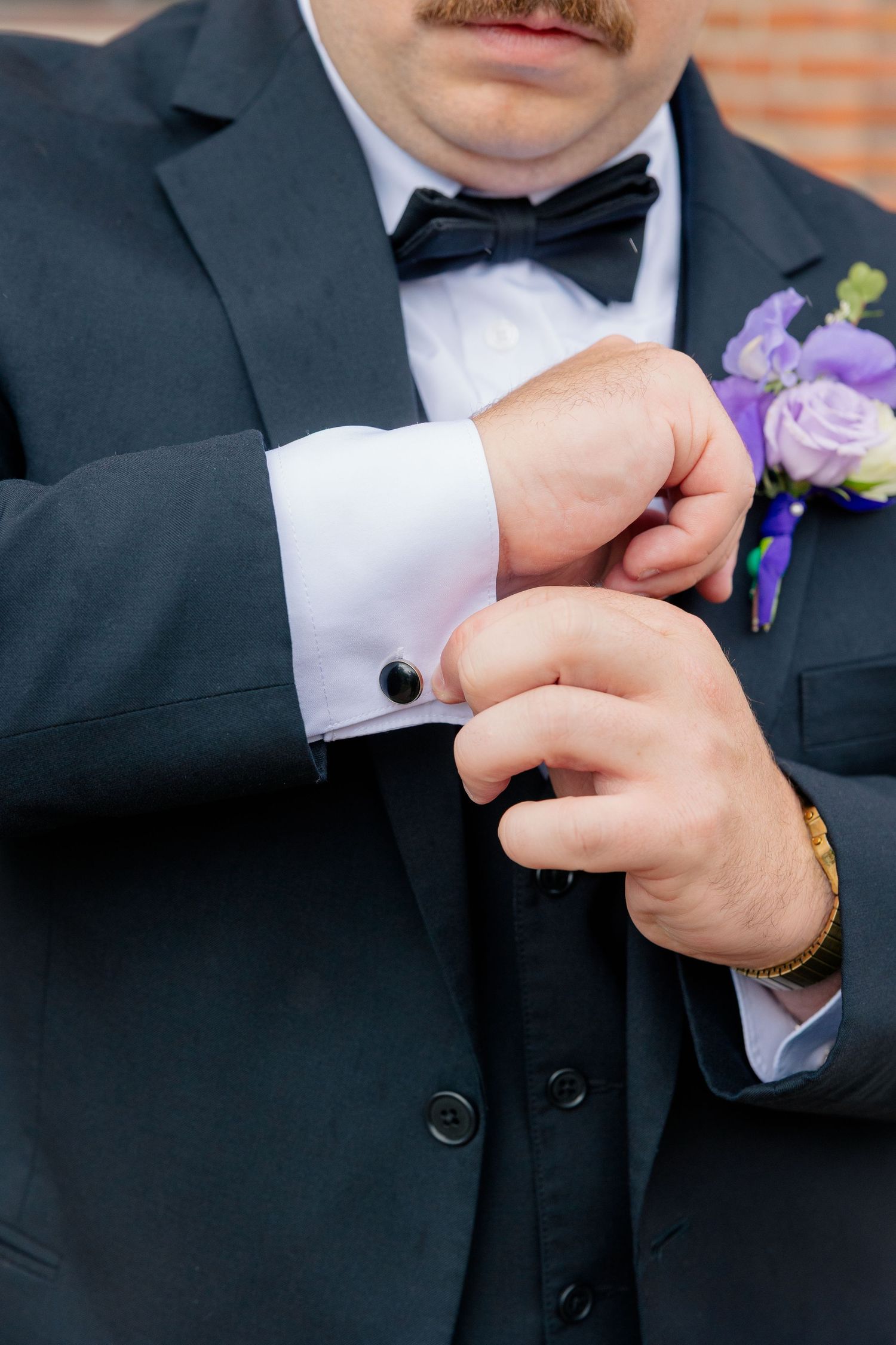 Groom in black tuxedo adjusting cufflinks with purple boutonniere on lapel.