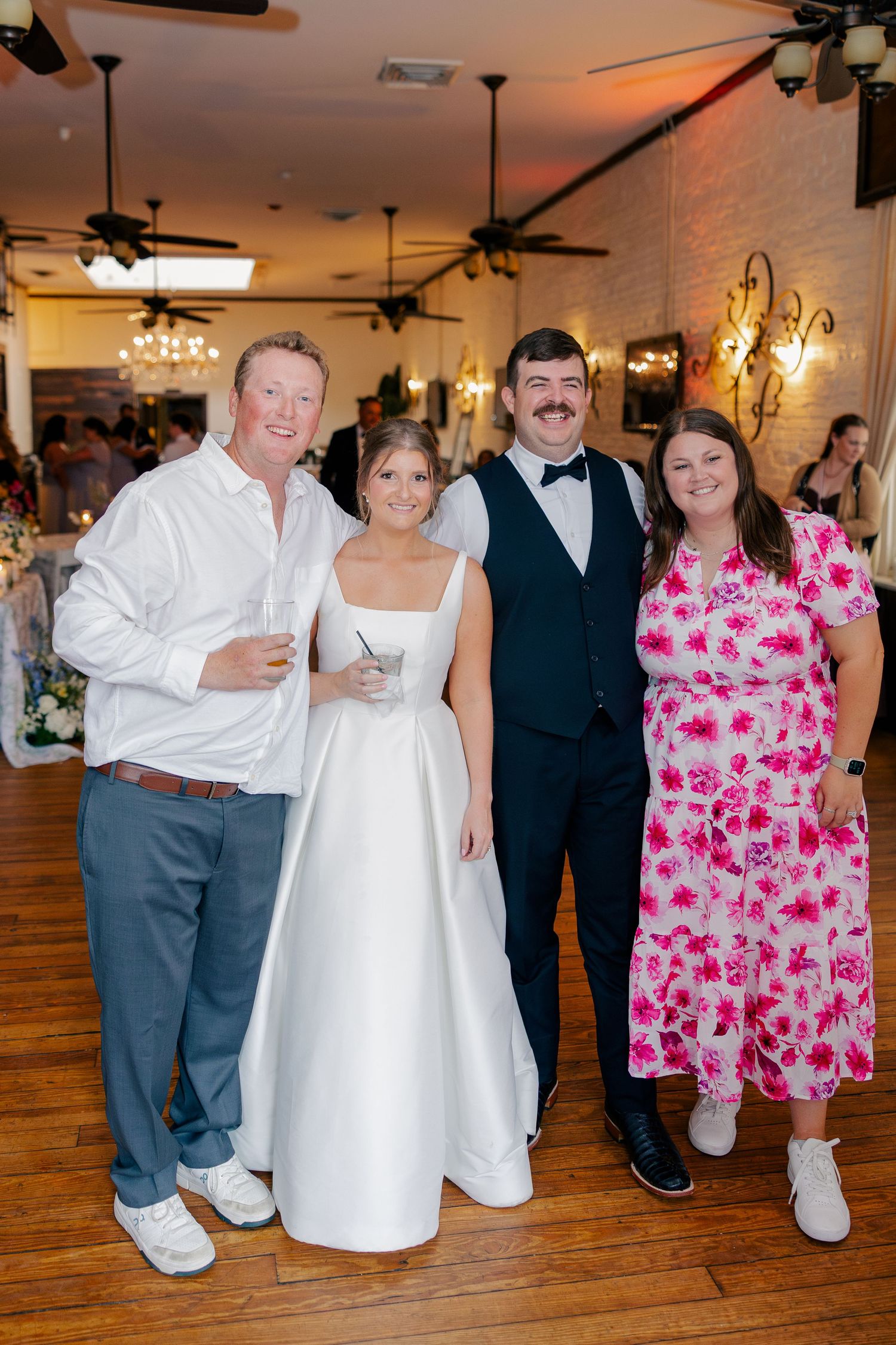 Bride in white gown and groom in navy tuxedo posing with two wedding guests on dance floor.