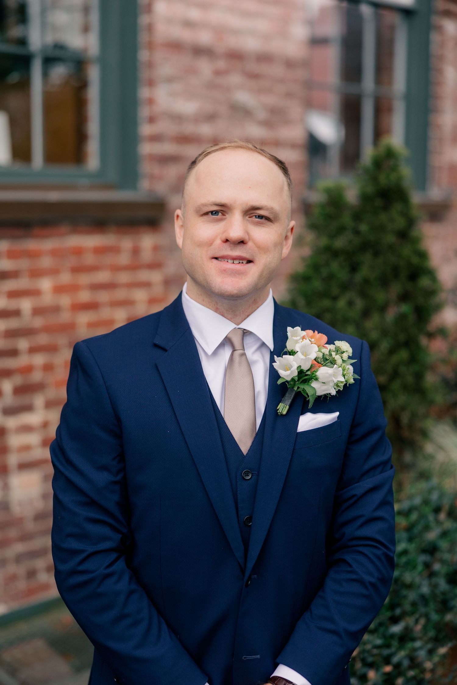 Groom in navy three-piece suit with floral boutonniere smiles outside brick building on wedding day.