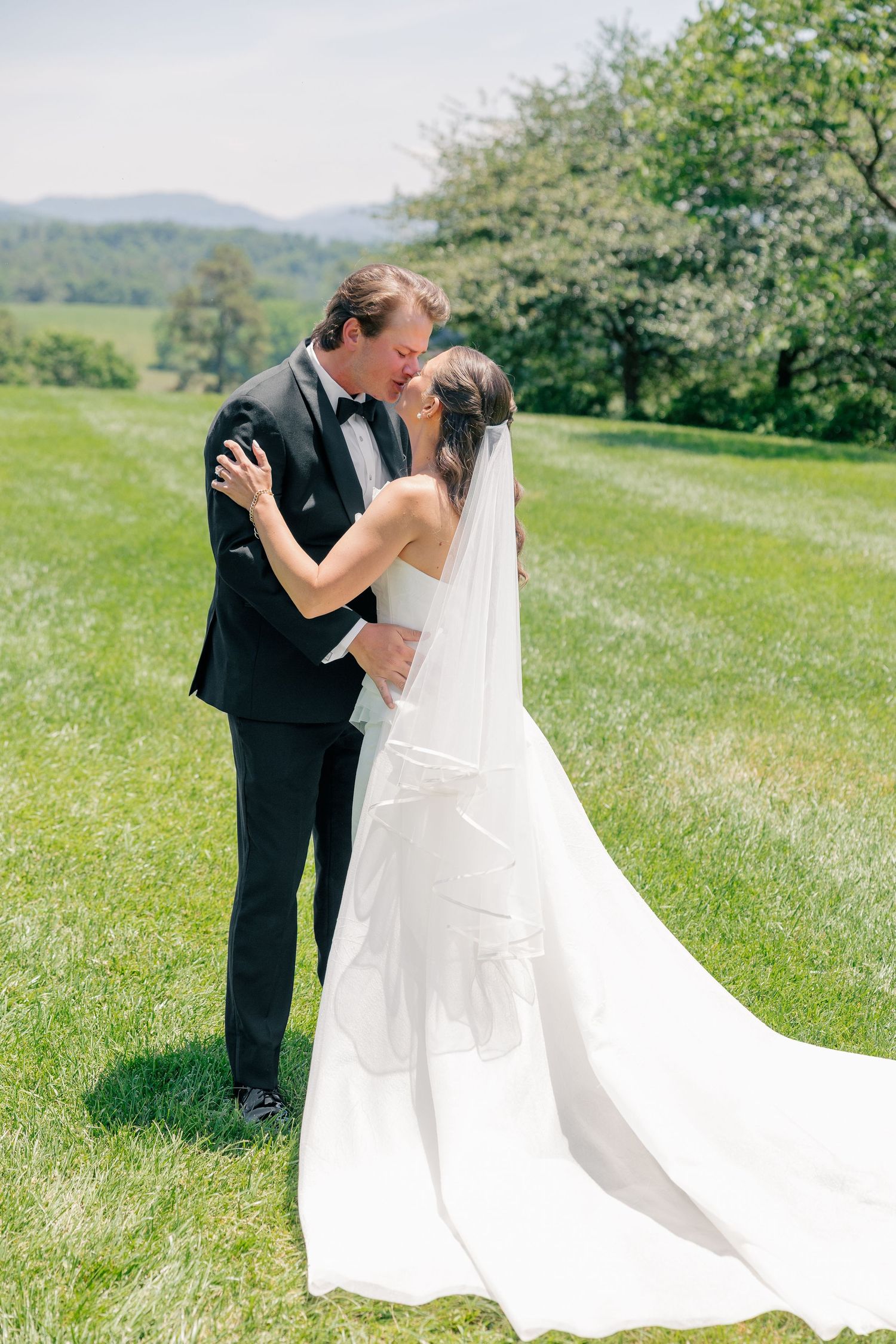 Bride and groom share a romantic kiss on a lush green lawn with mountain views behind them.