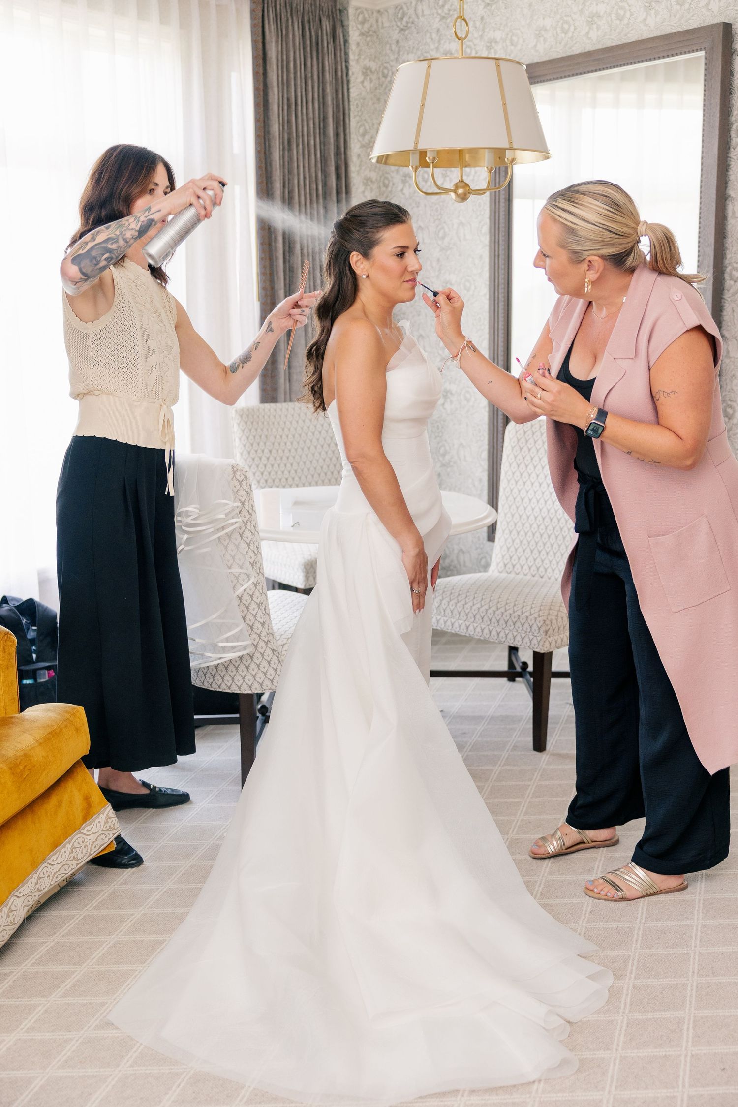 Bride gets final beauty touches from two stylists in a bright hotel suite before her wedding ceremony.