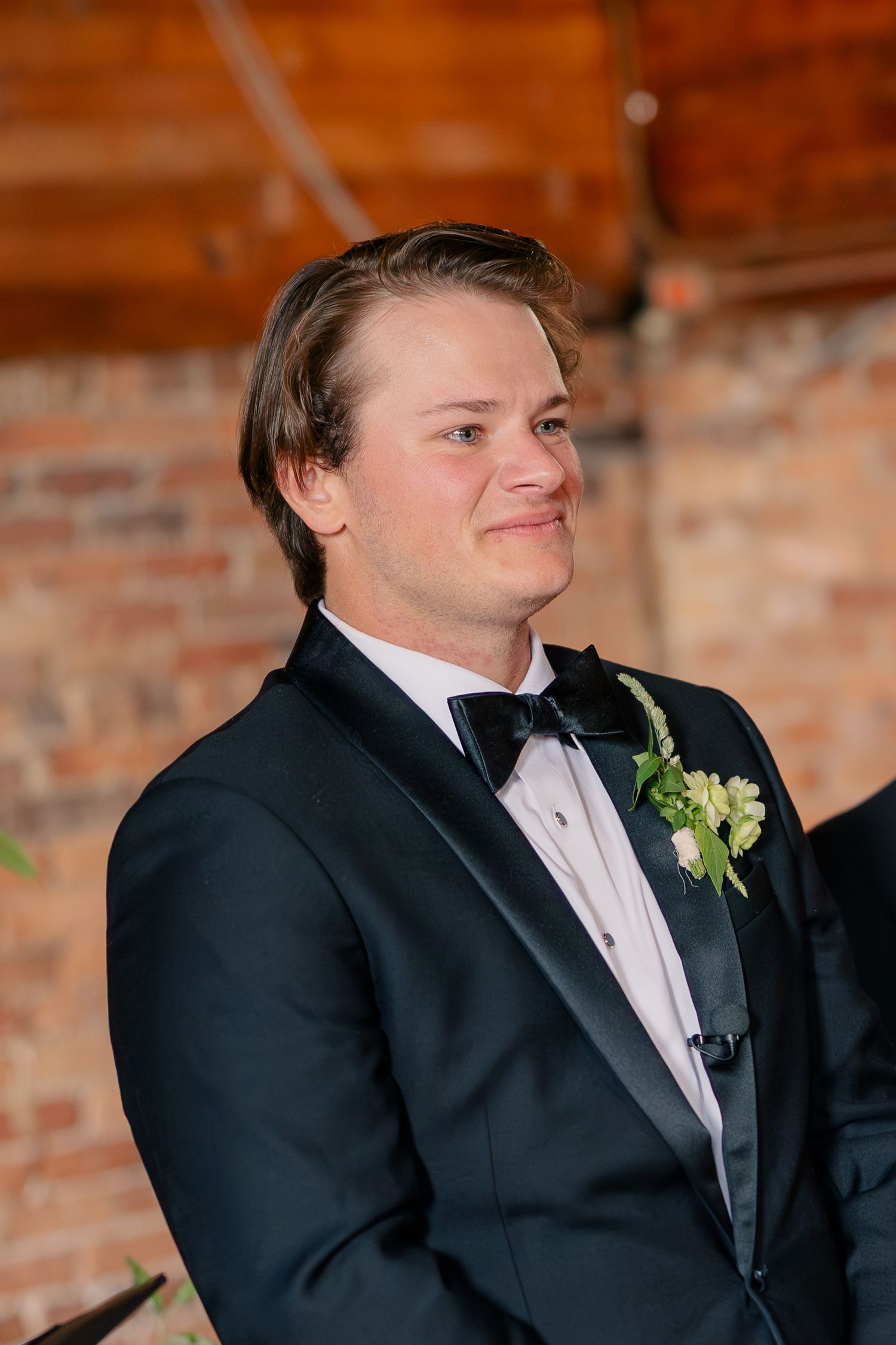 Groom smiles warmly in black tuxedo with floral boutonniere against rustic brick wall backdrop.