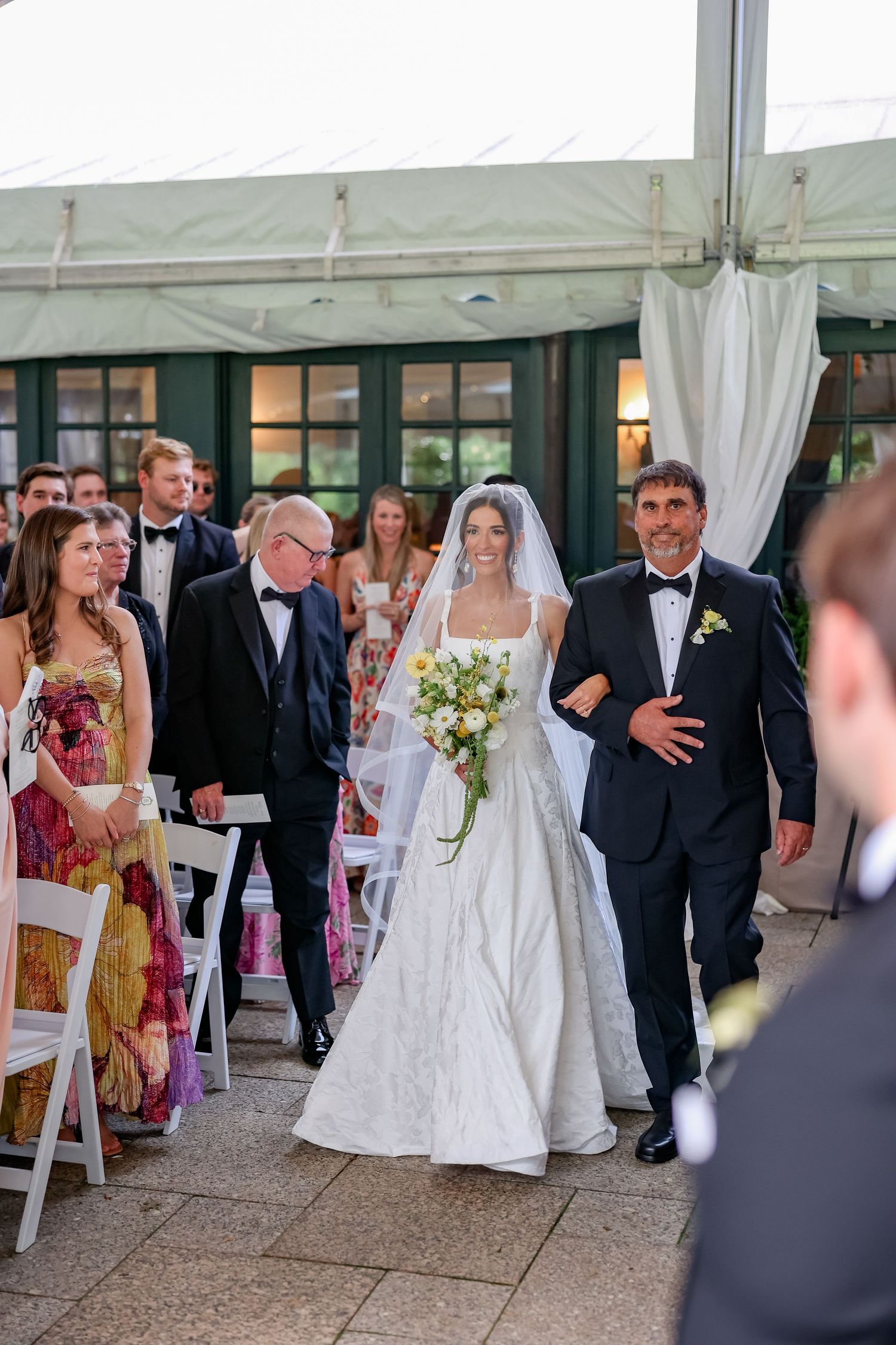 Bride walks down the aisle in a white gown and veil, holding a bouquet, escorted by a man in a tuxedo.