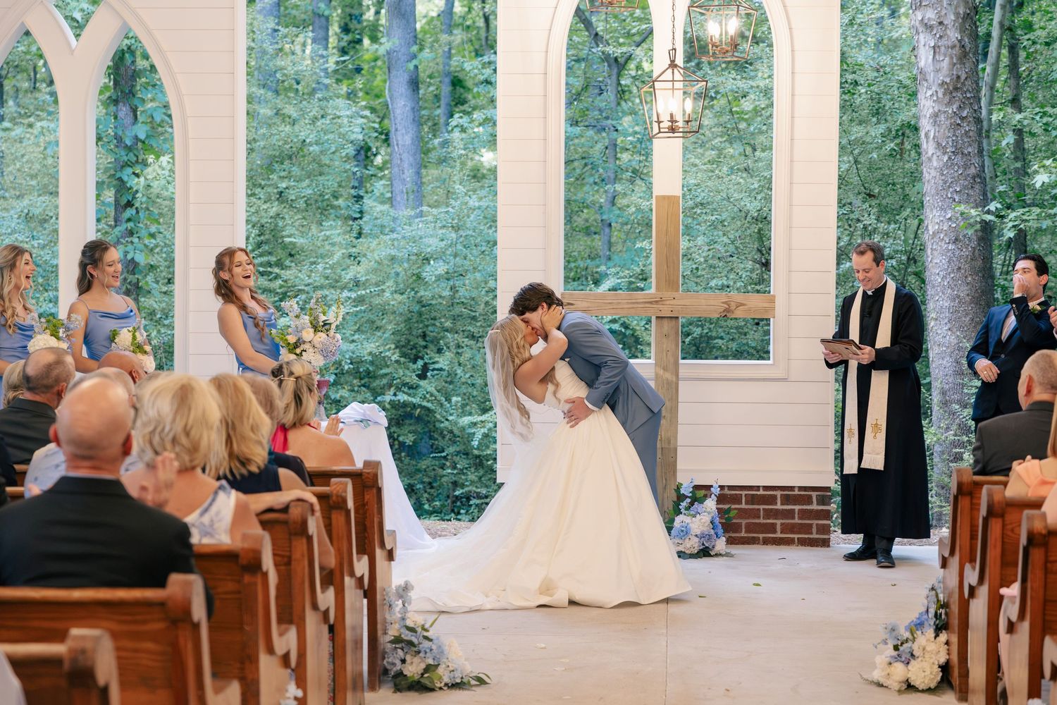 Bride and groom share first kiss at outdoor chapel wedding ceremony with bridesmaids and officiant nearby.