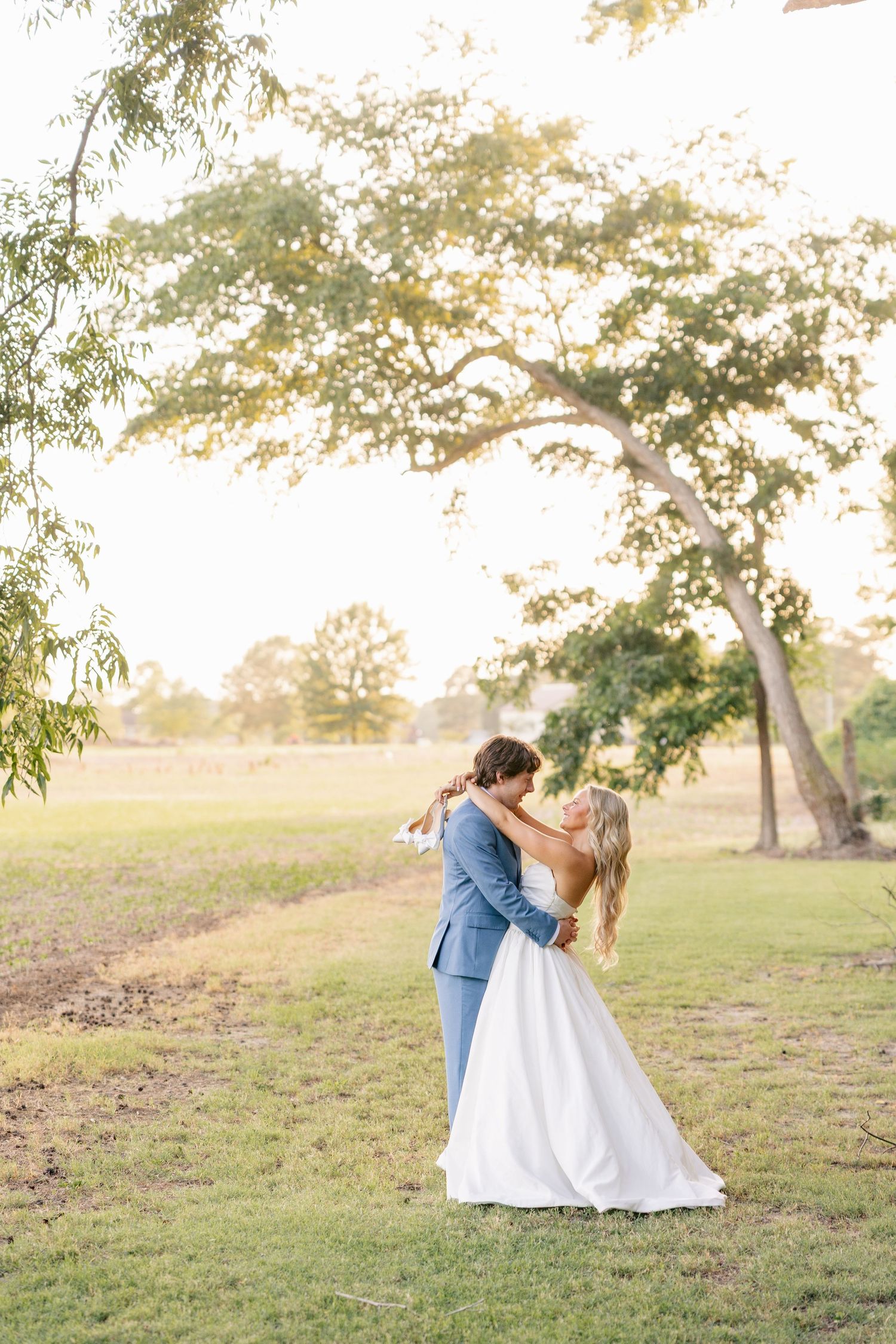 Bride and groom sharing a romantic kiss while embracing in a sunlit green field with trees in the background.