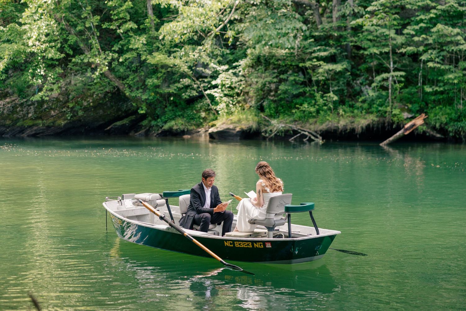 Bride and groom rowing a small green boat on a serene emerald green lake surrounded by lush trees.