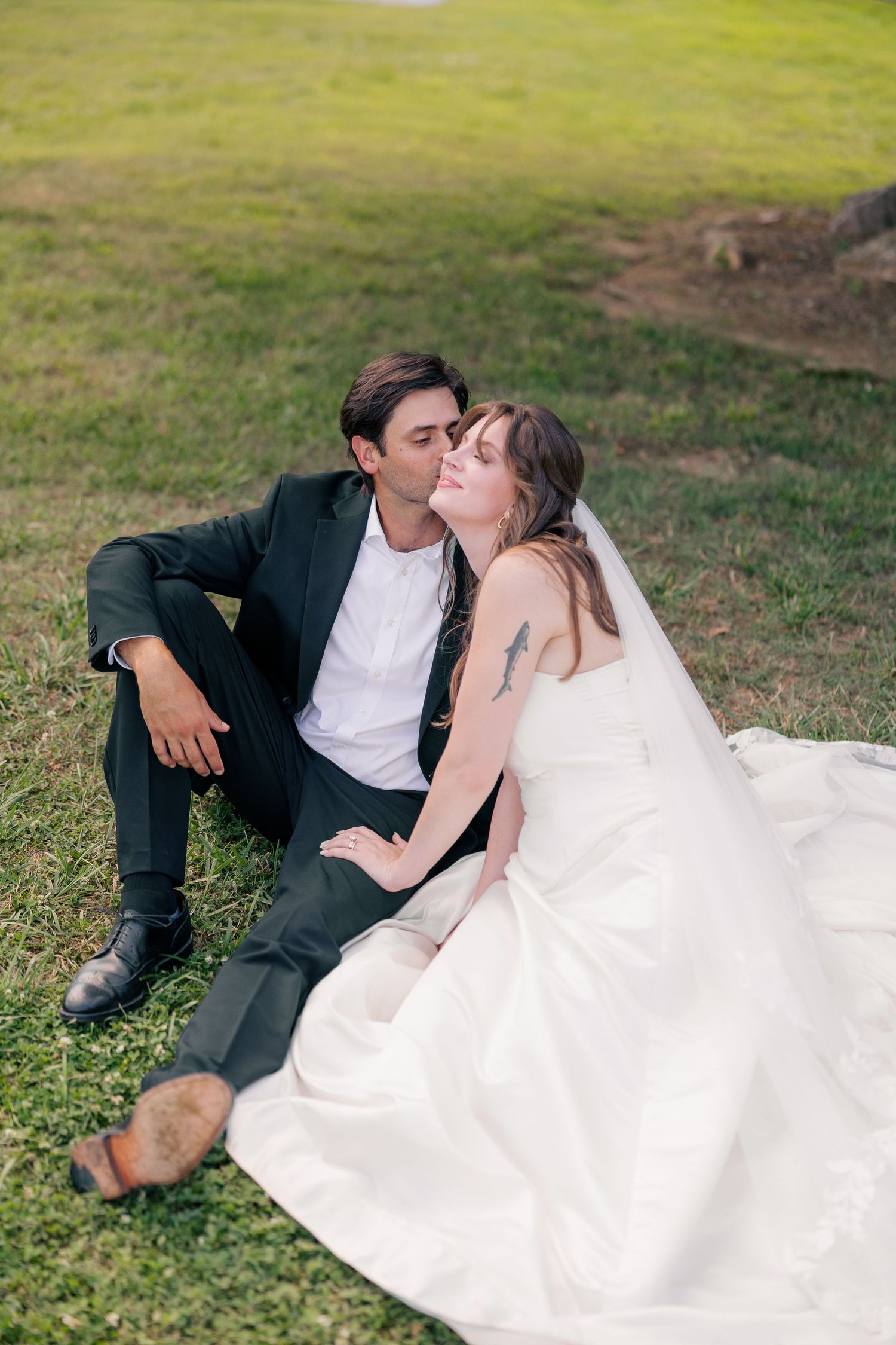 Bride and groom sitting on green grass, sharing a romantic kiss in their wedding attire outdoors.