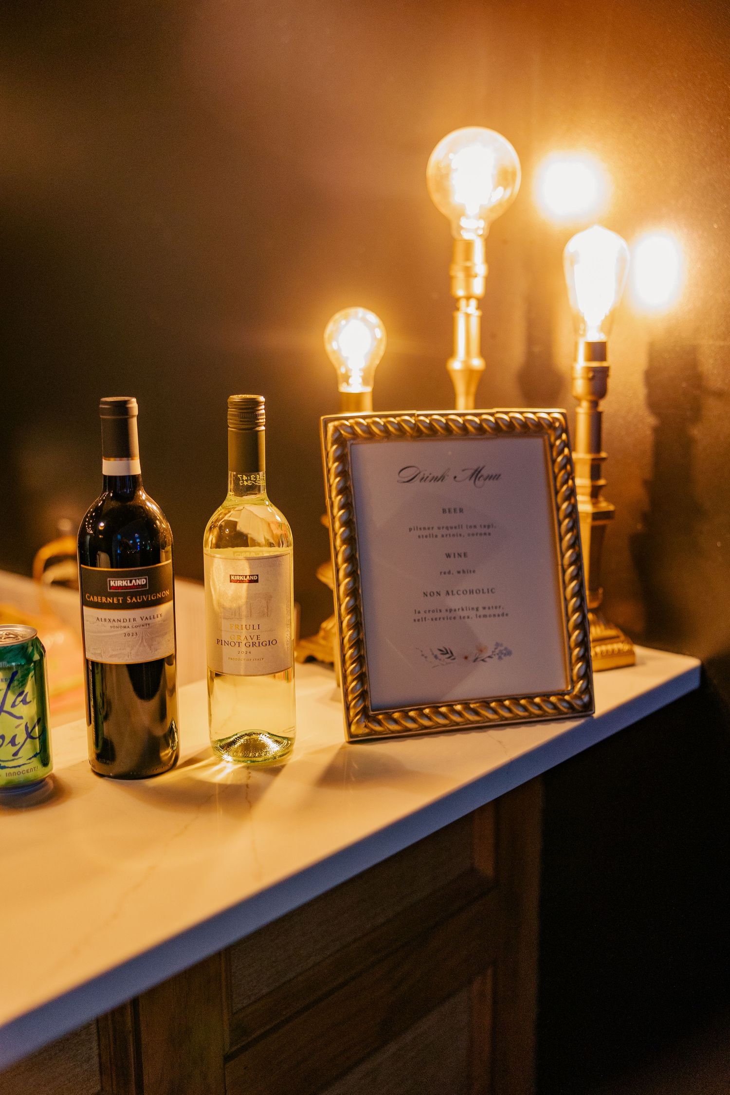 Wedding bar setup with wine bottles, framed drink menu, and vintage Edison bulb lamps in warm light.