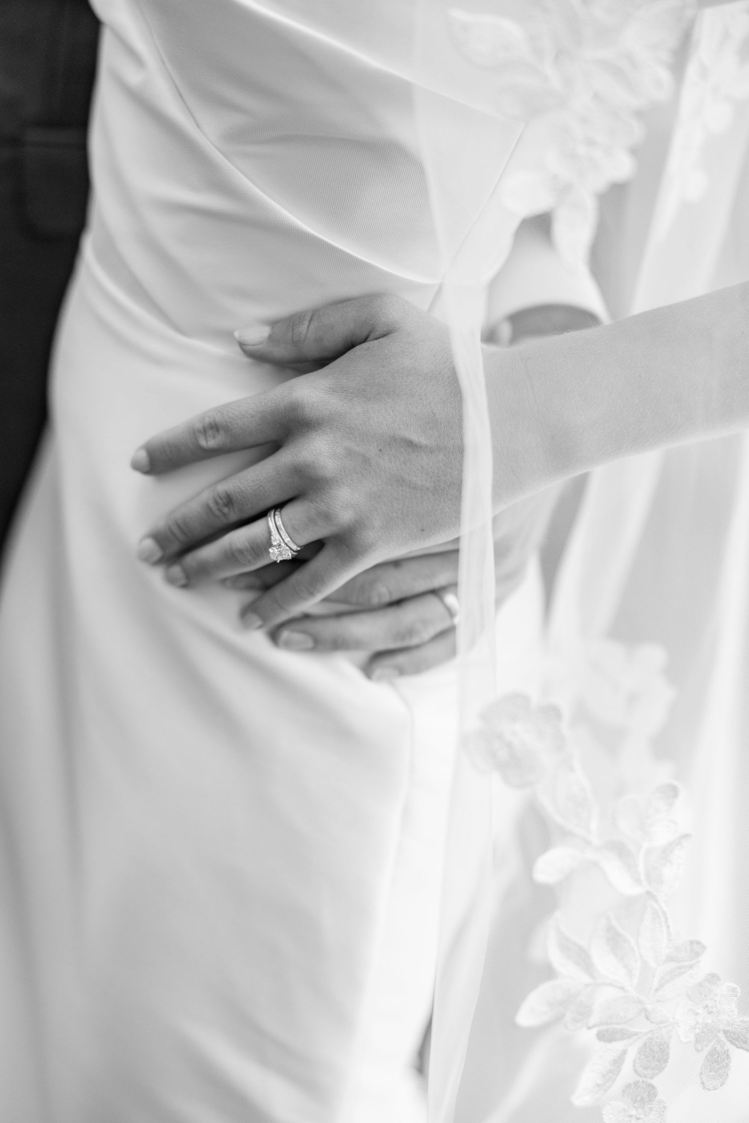 Black and white close-up of bride's hands showing diamond engagement ring on wedding dress.