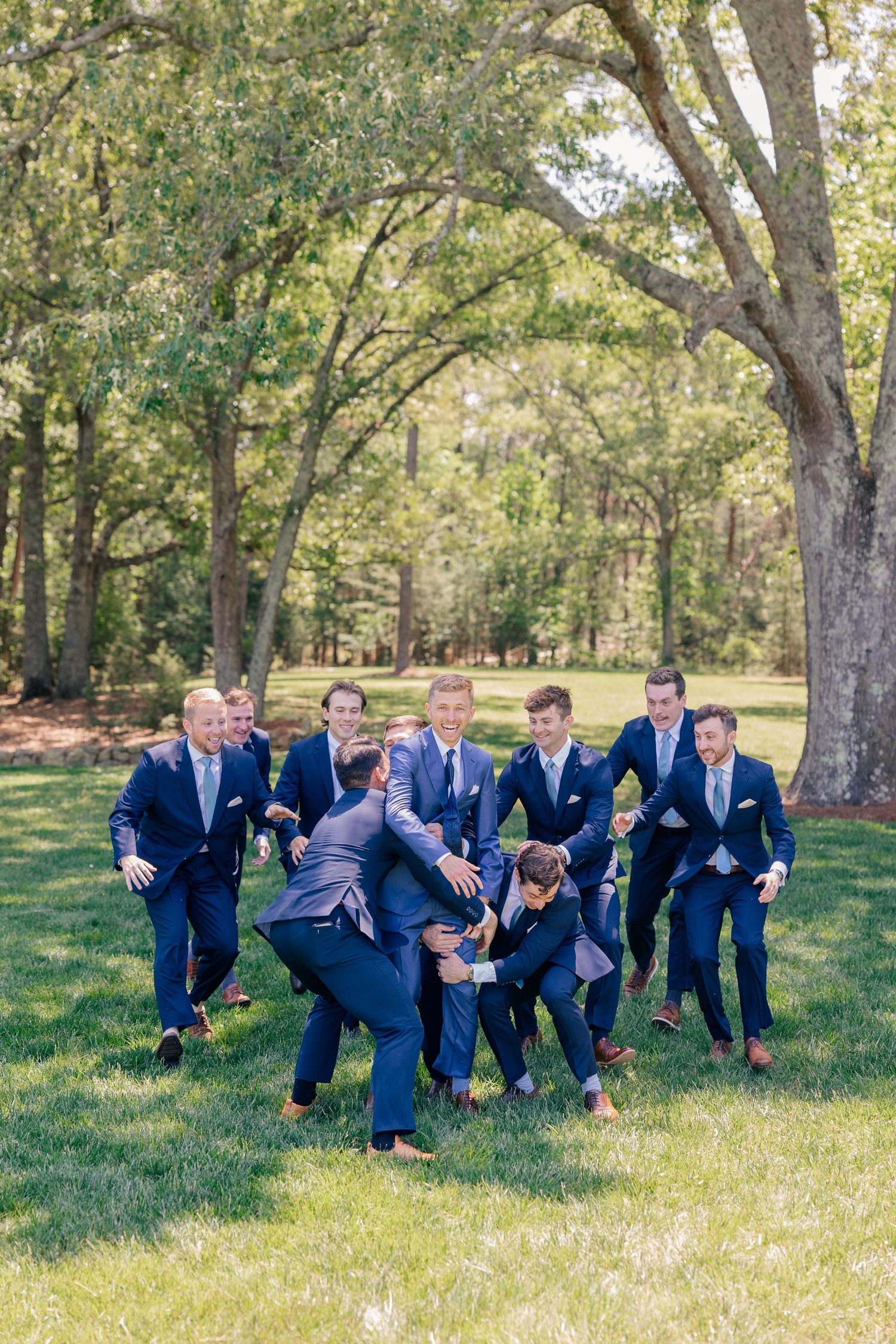 Groomsmen in navy suits playfully tackle each other on green lawn with trees in background at outdoor wedding.