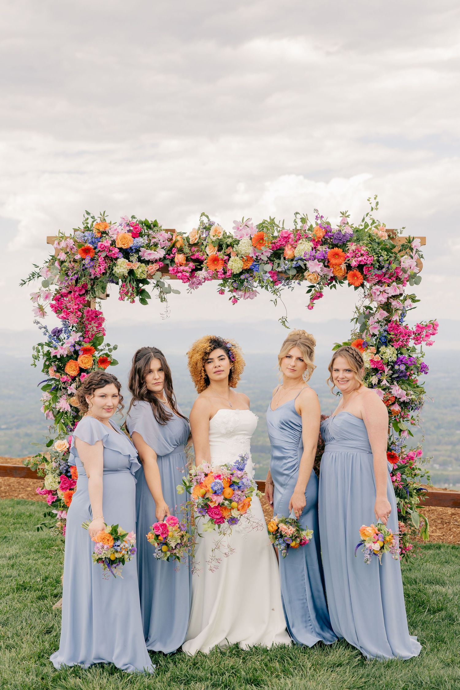 Bride and four bridesmaids in blue gowns pose under a colorful floral arch with mountain views.