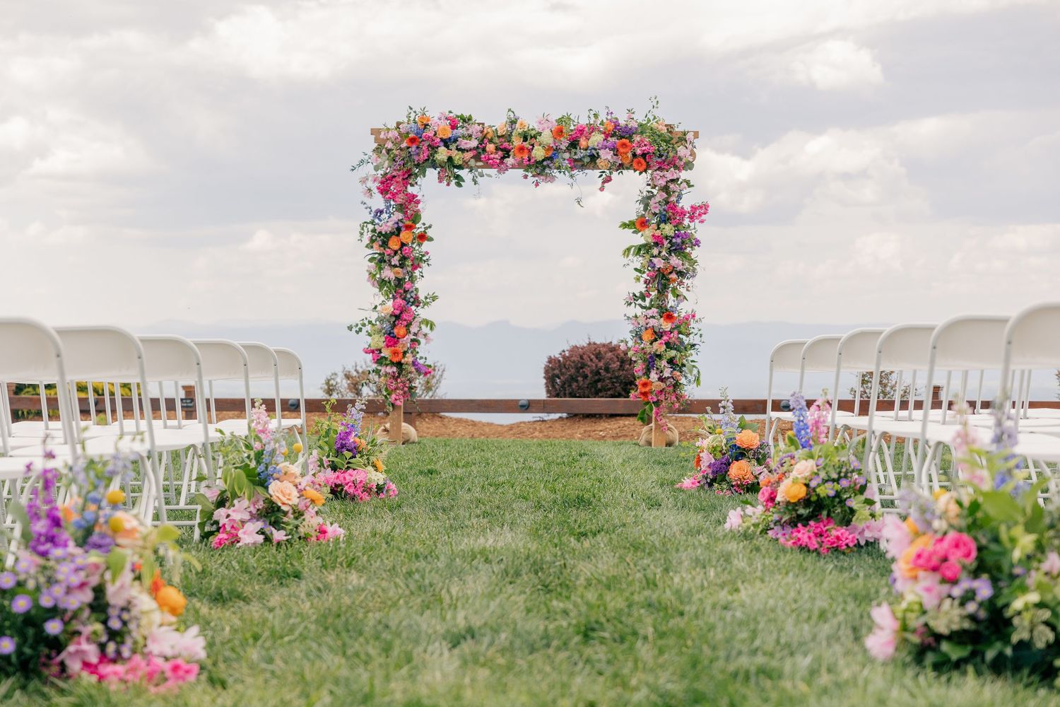 Colorful floral wedding arch frames outdoor aisle with white chairs and vibrant flower arrangements under cloudy sky.