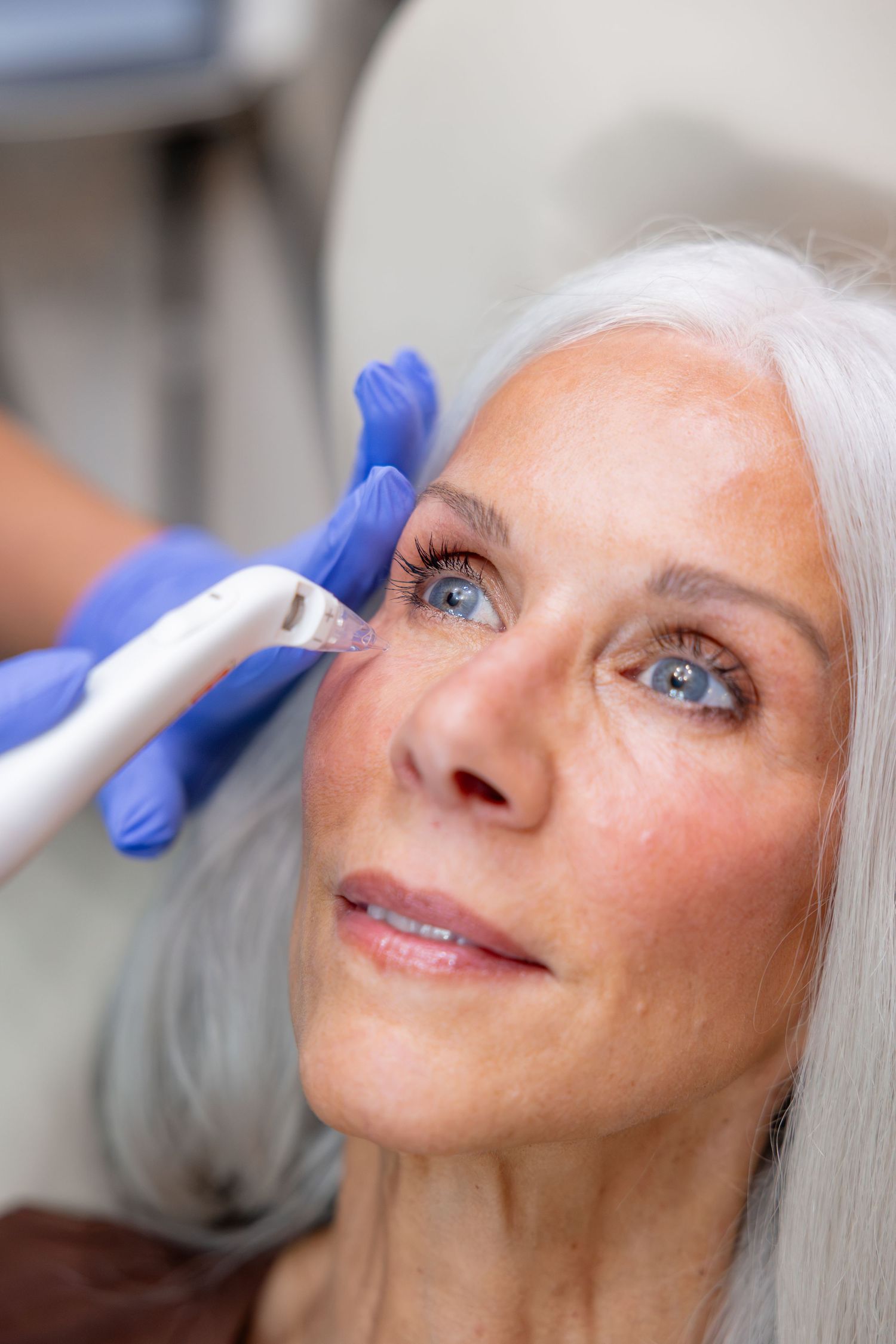 Older woman with gray hair receiving facial treatment with handheld device by gloved professional in clinic.