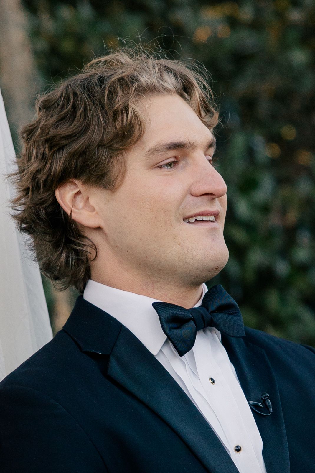 A person in formal attire wearing a black tuxedo and bow tie during what appears to be a special event or wedding.