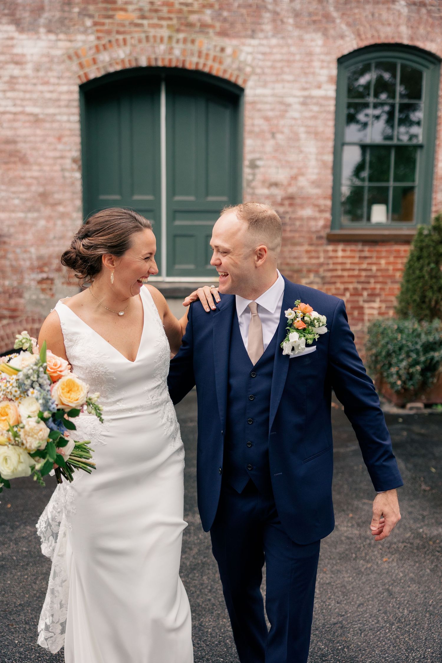 Happy bride and groom walk together laughing outside rustic brick building on their wedding day.