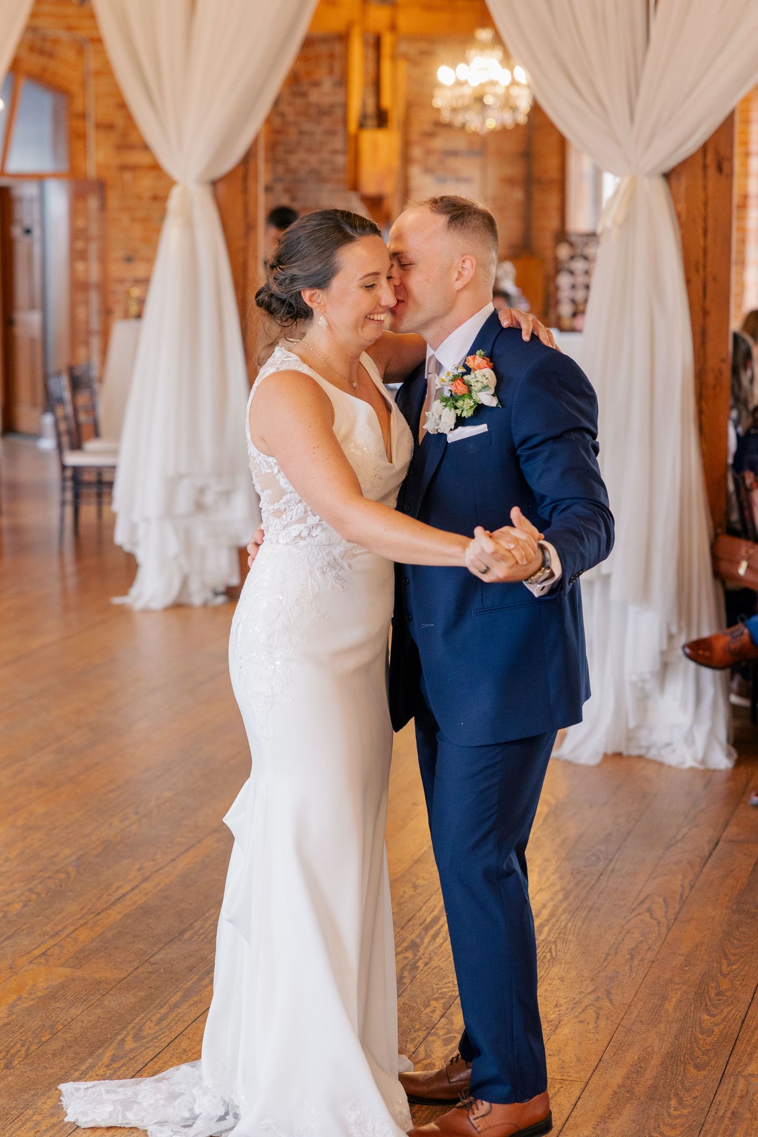 Bride and groom share an intimate first dance embrace in a romantic rustic brick reception hall with chandeliers.