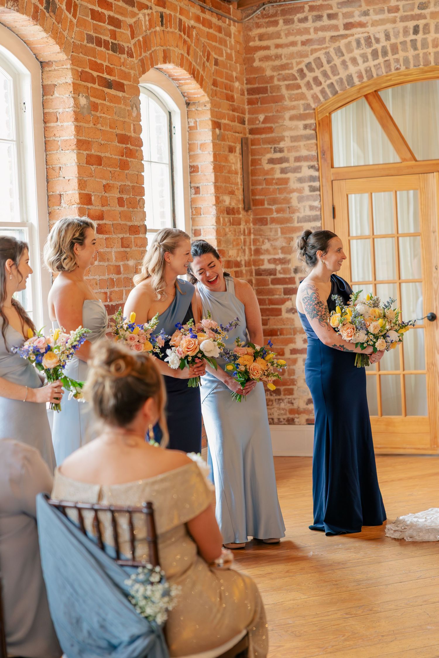 Bridesmaids in blue gowns holding colorful bouquets laugh together in sunlit brick venue.