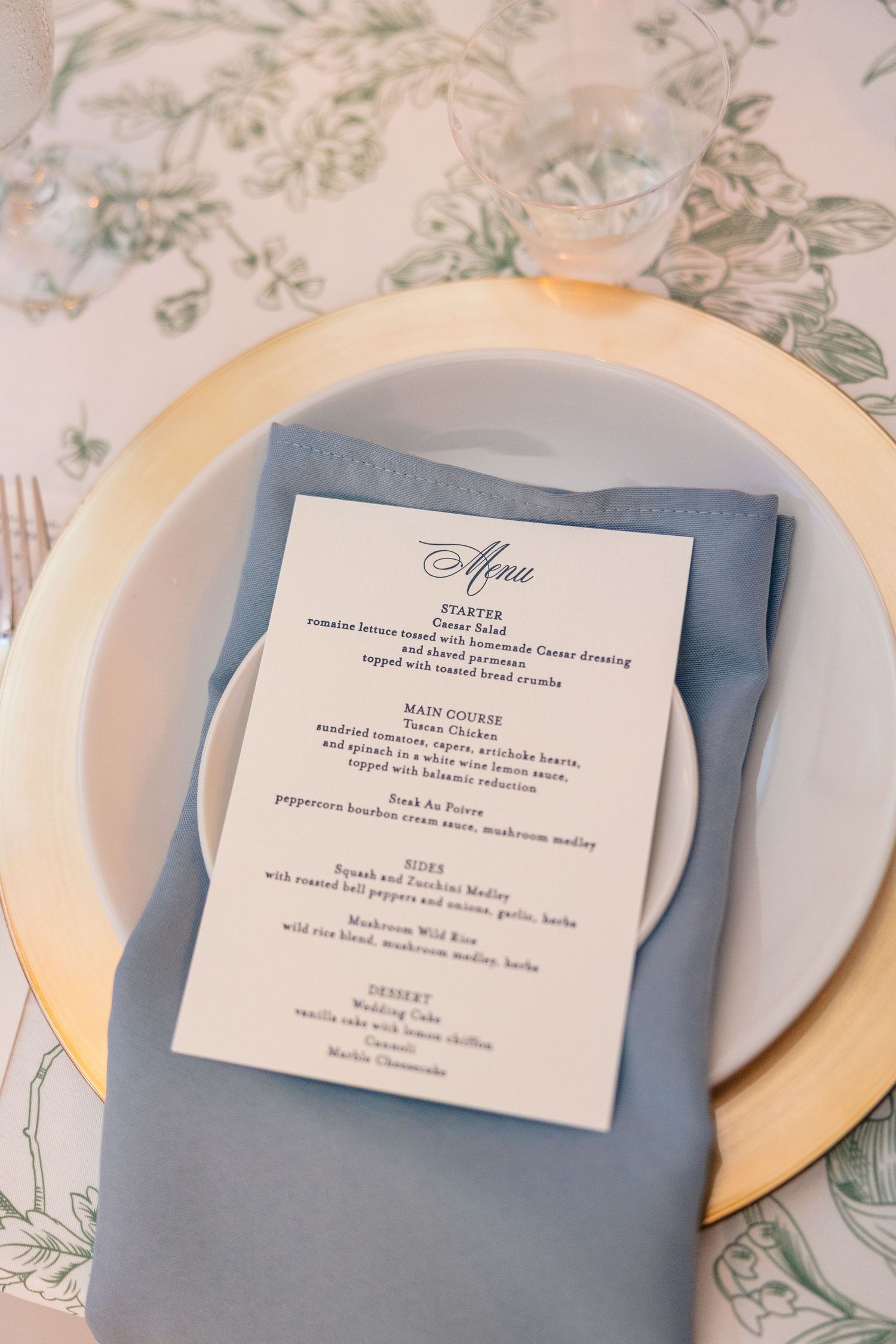 Elegant wedding place setting with gold charger, blue napkin, and printed menu card on floral tablecloth.