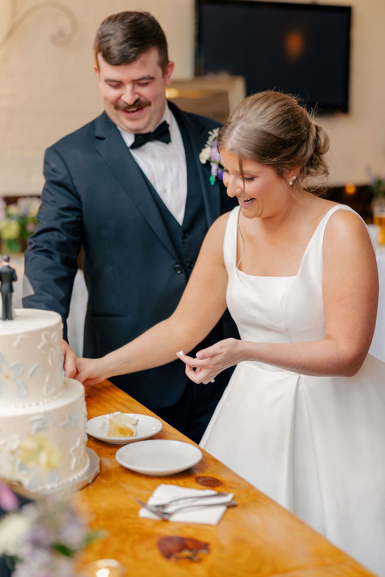 Smiling bride and groom cut elegant white wedding cake together at rustic wood reception table.
