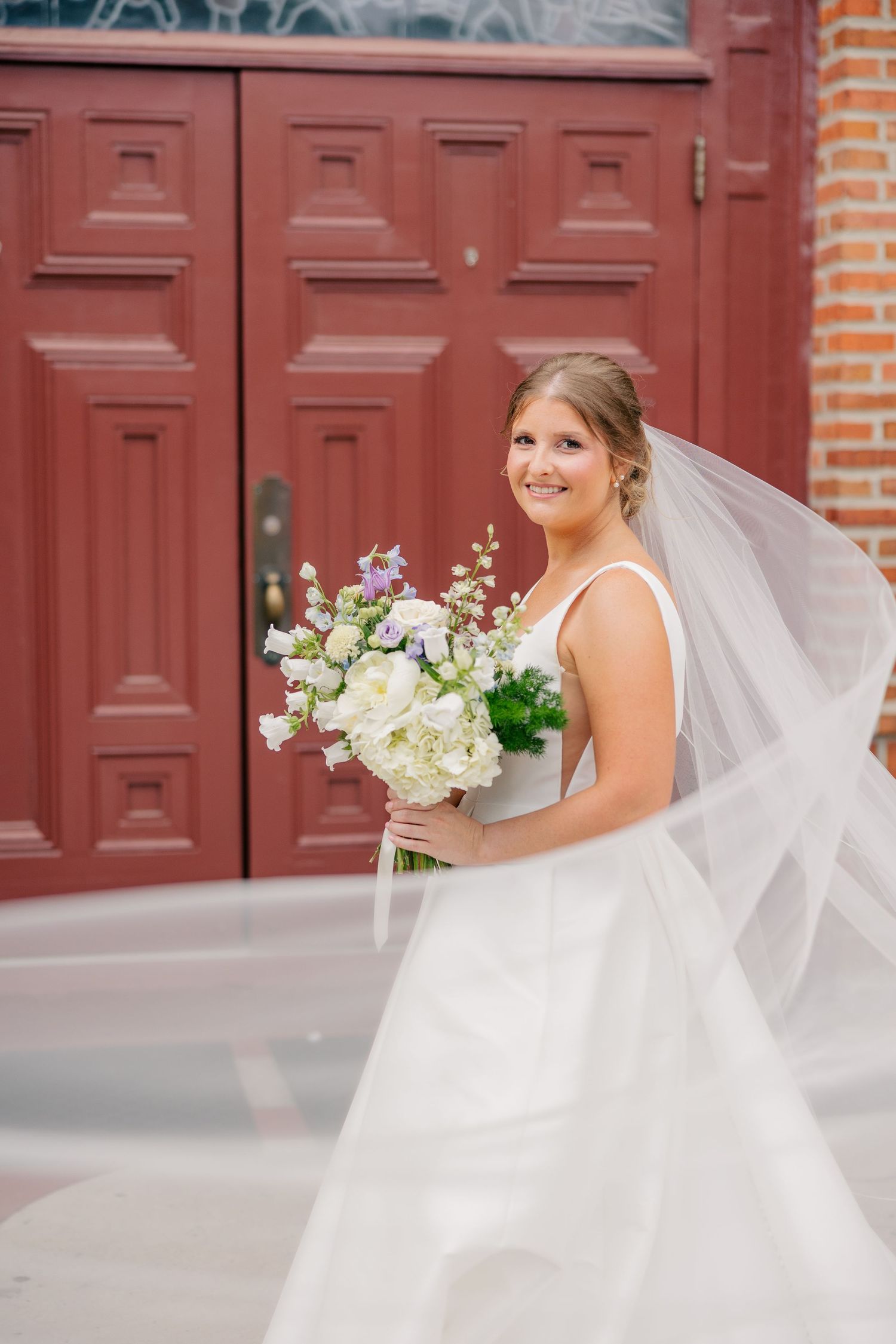 Smiling bride in white gown holding a white and blue floral bouquet in front of red wooden doors.