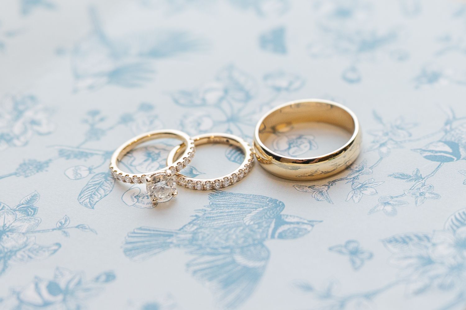 Three gold wedding rings with diamonds on a blue floral background, including an engagement ring and two bands.