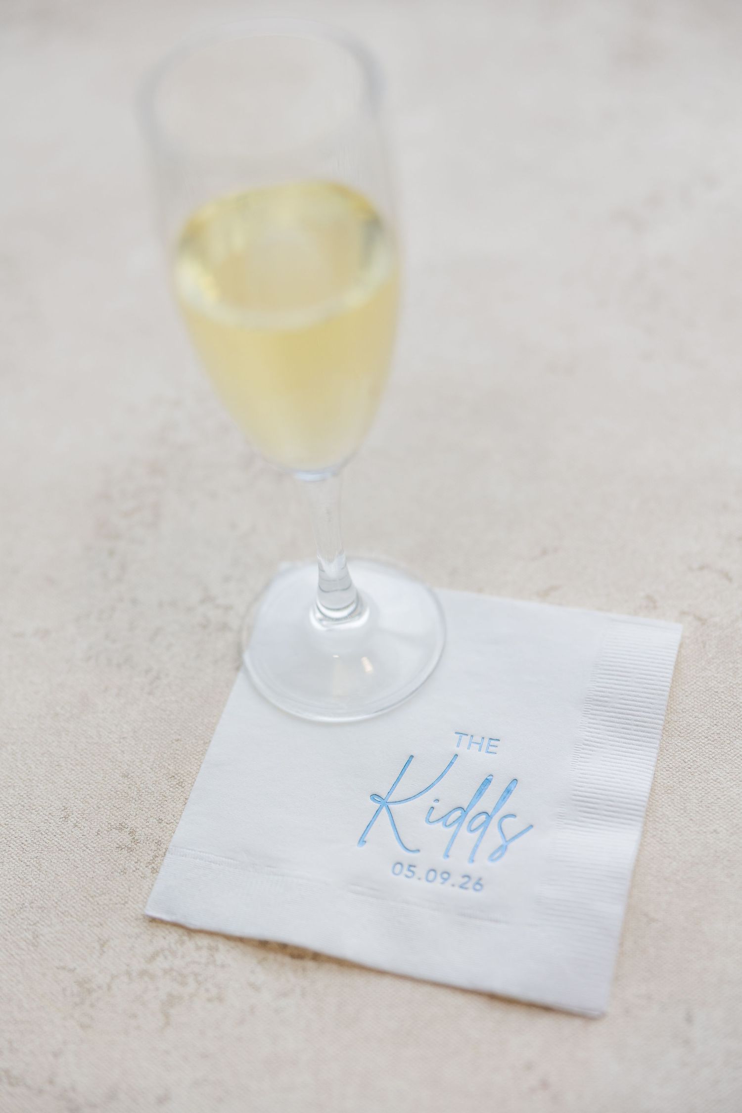 Personalized white wedding napkin reading The Kidds with date beside a champagne flute at a reception.