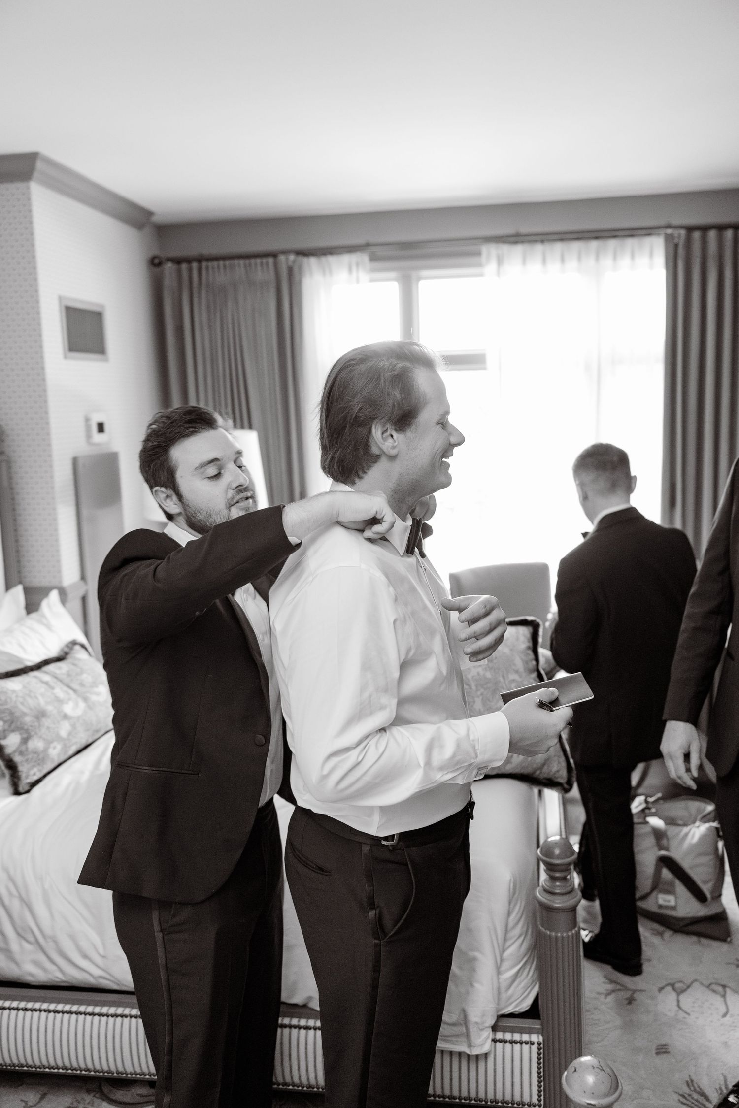 Black and white photo of groomsmen helping each other get dressed in a hotel room before the wedding ceremony.