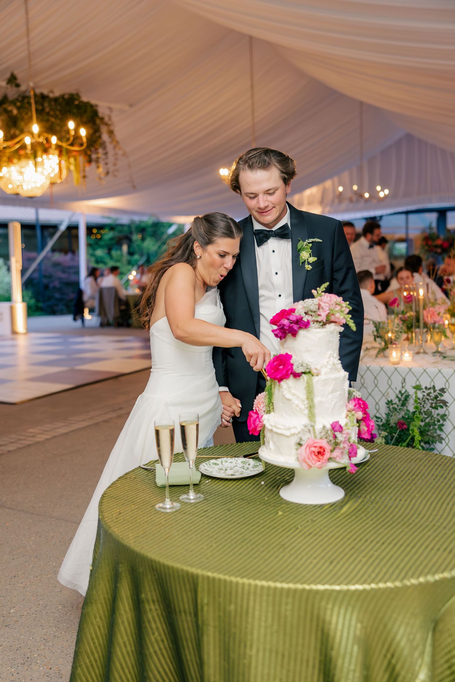 Bride and groom cutting their white wedding cake adorned with pink flowers at an elegant tented reception.