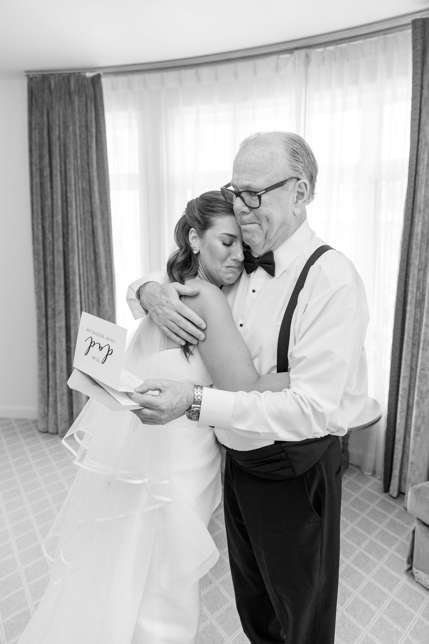 Black and white photo of a bride embracing an older man in suspenders and bow tie while reading a card.