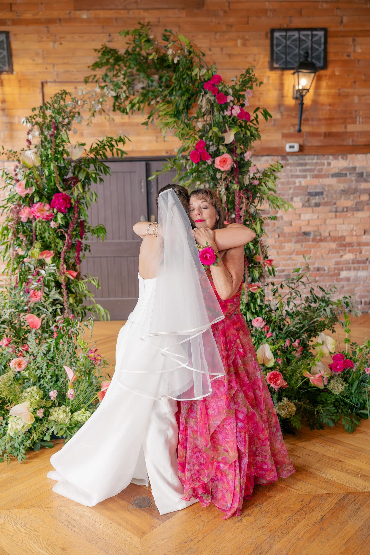 Bride and woman in pink floral dress share warm embrace before lush floral arch at wedding.
