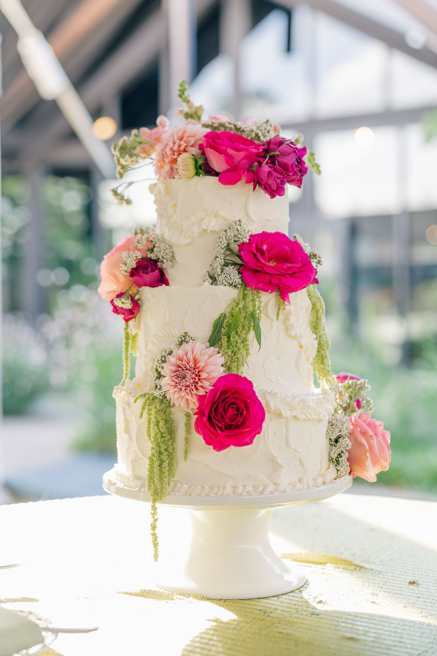 Three-tier white wedding cake adorned with fresh pink roses, dahlias, and cascading greenery.