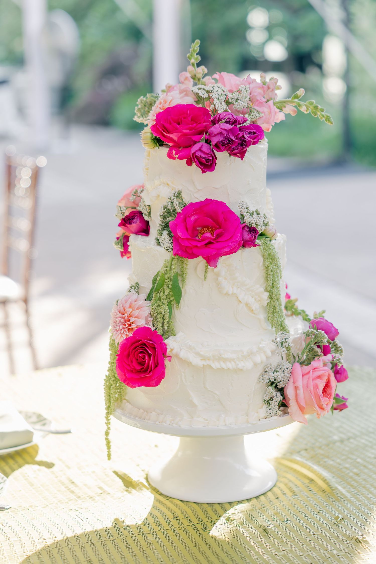 Elegant three-tier white wedding cake adorned with vibrant pink roses and greenery on a white pedestal stand.