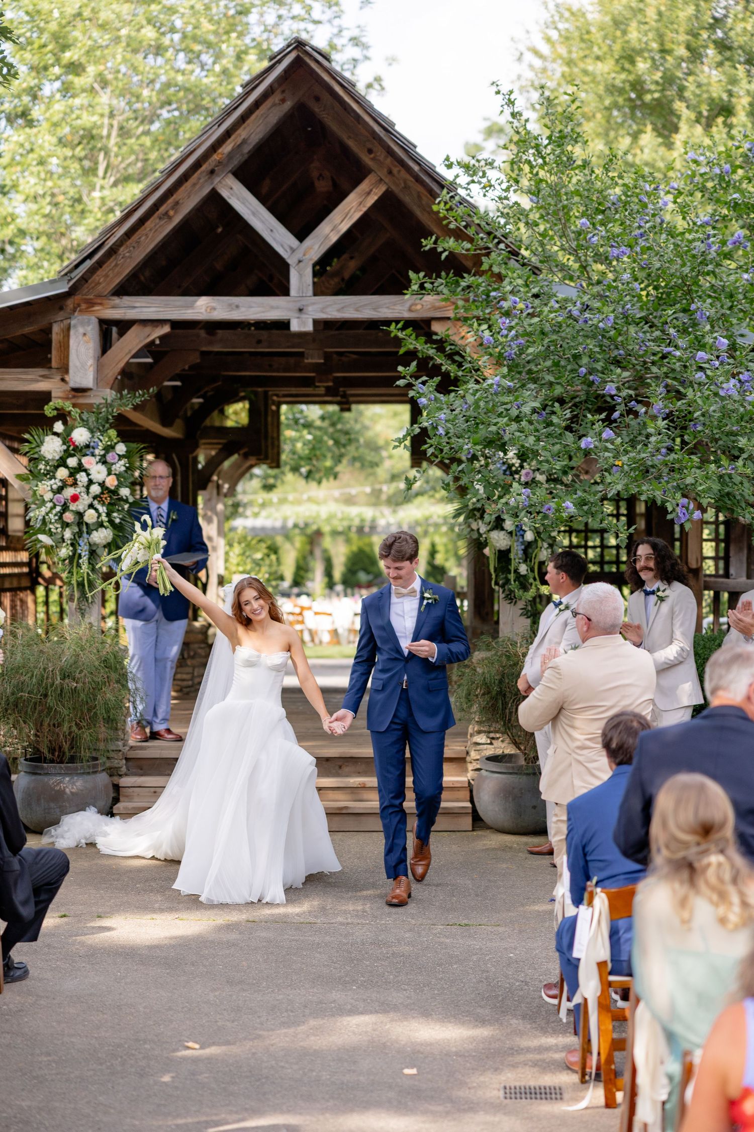 A couple dances under a rustic wooden pavilion surrounded by wedding guests and natural greenery.